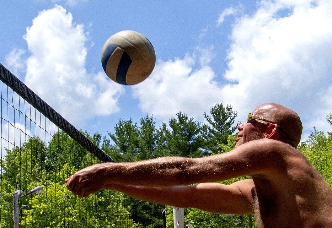 A volleyball player at White Thorn Lodge lunges forward with arms outstretched to bump the ball over the net during a sunny outdoor match, with trees and blue sky in the background. A volleyball player at White Thorn Lodge lunges forward with arms outstretched to bump the ball over the net during a sunny outdoor match, with trees and blue sky in the background.