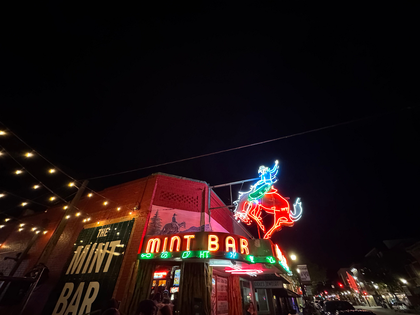 Neon sign of The Mint Bar in Wyoming at night, glowing with a red bucking horse and cowboy. String lights hang above, casting a warm glow. The old brick bar stands under a vast, dark sky, capturing the spirit of quiet Western grit and small-town life. Neon sign of The Mint Bar in Wyoming at night, glowing with a red bucking horse and cowboy. String lights hang above, casting a warm glow. The old brick bar stands under a vast, dark sky, capturing the spirit of quiet Western grit and small-town life.