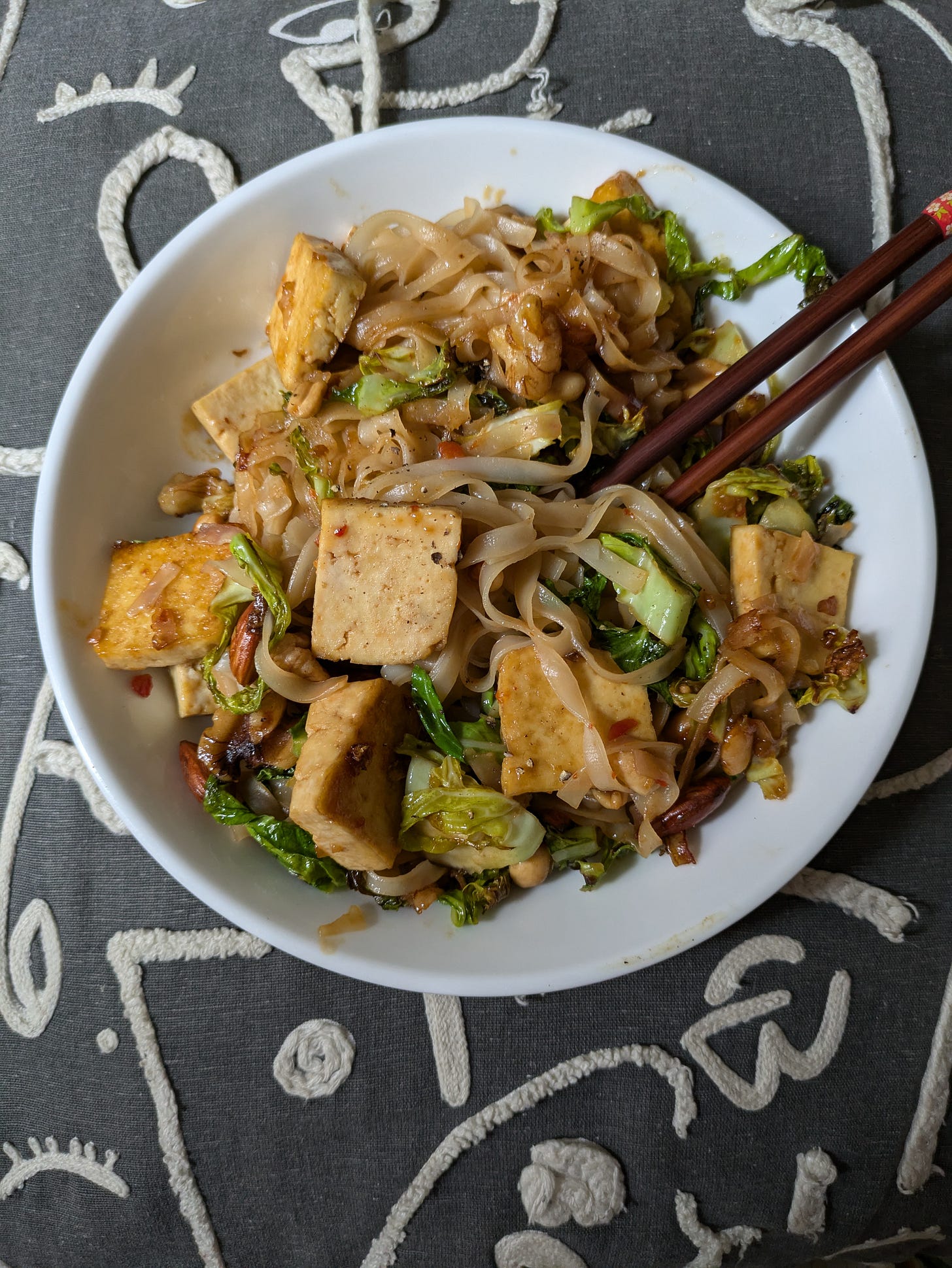 ID. A photo of a white plate of delicious stir fry - rice noodles, sauce, tofu, greens and almonds - with wooden chopsticks. ID. A photo of a white plate of delicious stir fry - rice noodles, sauce, tofu, greens and almonds - with wooden chopsticks.
