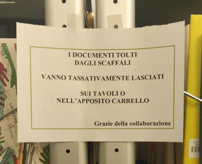 Un avviso stampato su un foglio bianco, con un bordo decorativo verde chiaro, è appeso a un ripiano di scaffale in una biblioteca o un archivio. Il messaggio in italiano, scritto in maiuscolo, ricorda agli utenti: "I DOCUMENTI TOLTI DAGLI SCAFFALI VANNO TASSATIVAMENTE LASCIATI SUI TAVOLI O NELL'APPOSITO CARRELLO". Sotto, si legge "Grazie della collaborazione". Lo sfondo mostra altri scaffali e il dorso di un libro o faldone giallo sulla destra.