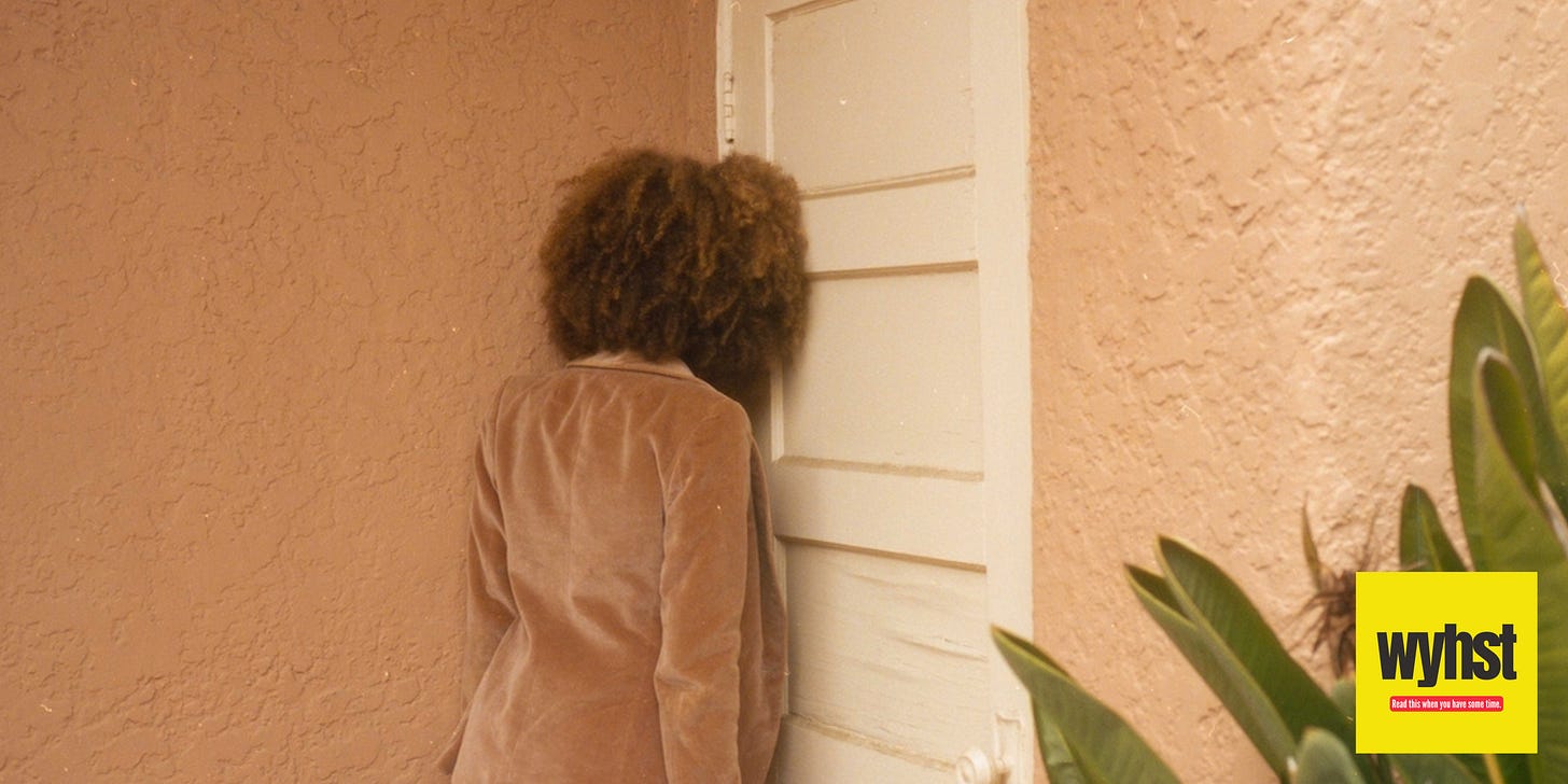 A woman with an afro and wearing a brown jacket has her back facing the camera and is leaning her head against the wall.