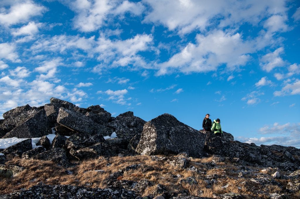 Two hikers sit on a jumble of lichen-covered granite blocks on a high ridge under a wide blue sky scattered with clouds, brown tundra in the foreground.