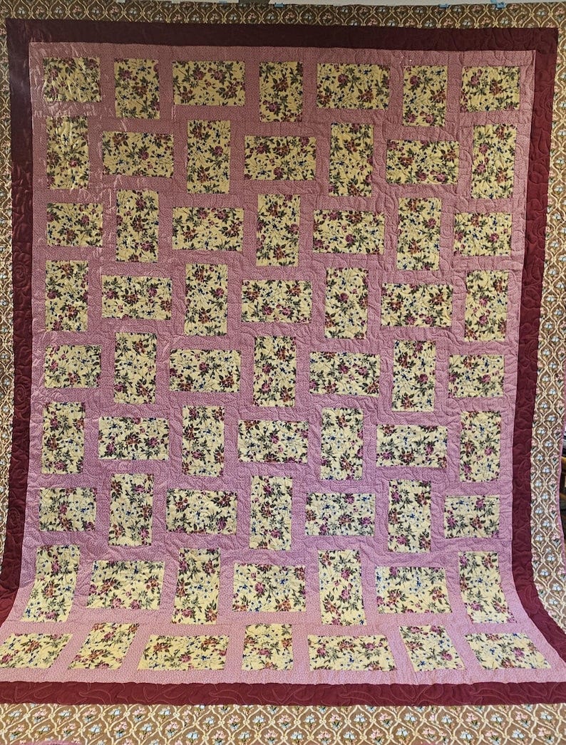May include: A handmade quilt featuring a repeating pattern of floral blocks in shades of yellow, pink, and burgundy. The blocks are set against a pink background, with a burgundy border and trim. May include: A handmade quilt featuring a repeating pattern of floral blocks in shades of yellow, pink, and burgundy. The blocks are set against a pink background, with a burgundy border and trim.