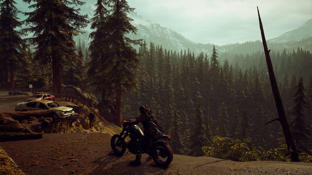 Still image from Days Gone featuring Deacon, a male video game character on a motorbike, facing into a dense pine woodland valley below a broken road