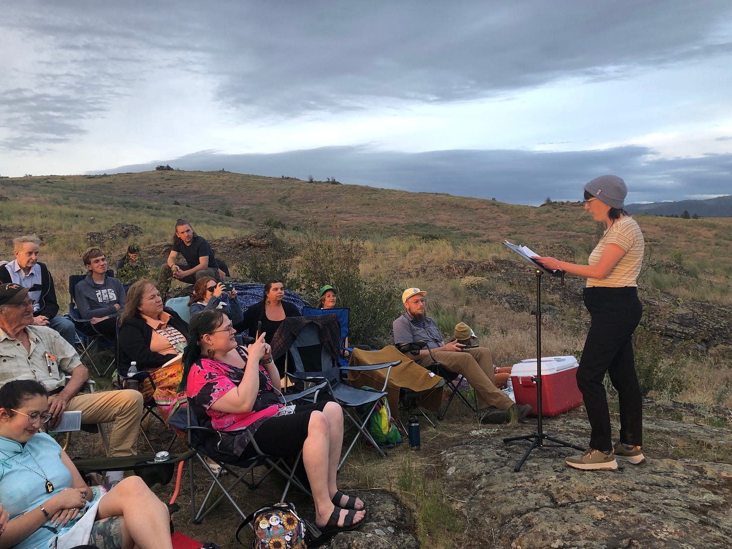 A nonbinary person reading to a small crowd of people outdoors in a rocky area of dried grass.