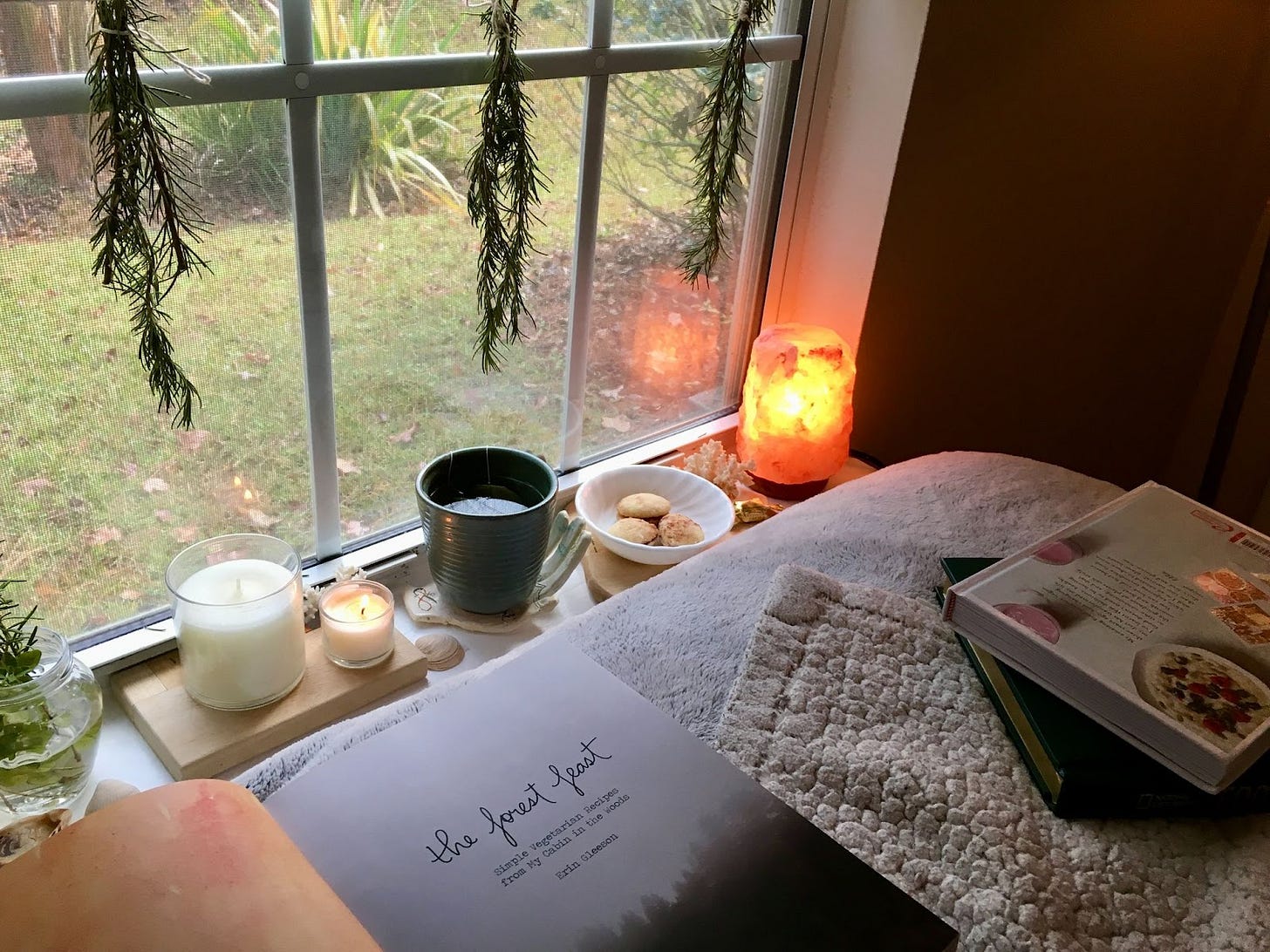 A cozy window scene: drying rosemary bundles hang over a windowsill with tea, candles, and a salt lamp. Books and blankets in the foreground.