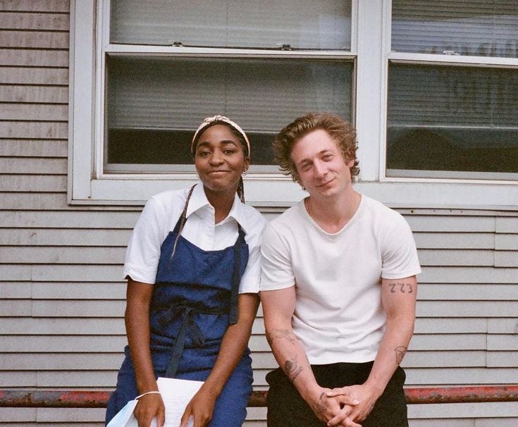 This may contain: two people sitting on a bench in front of a house and one person is wearing an apron