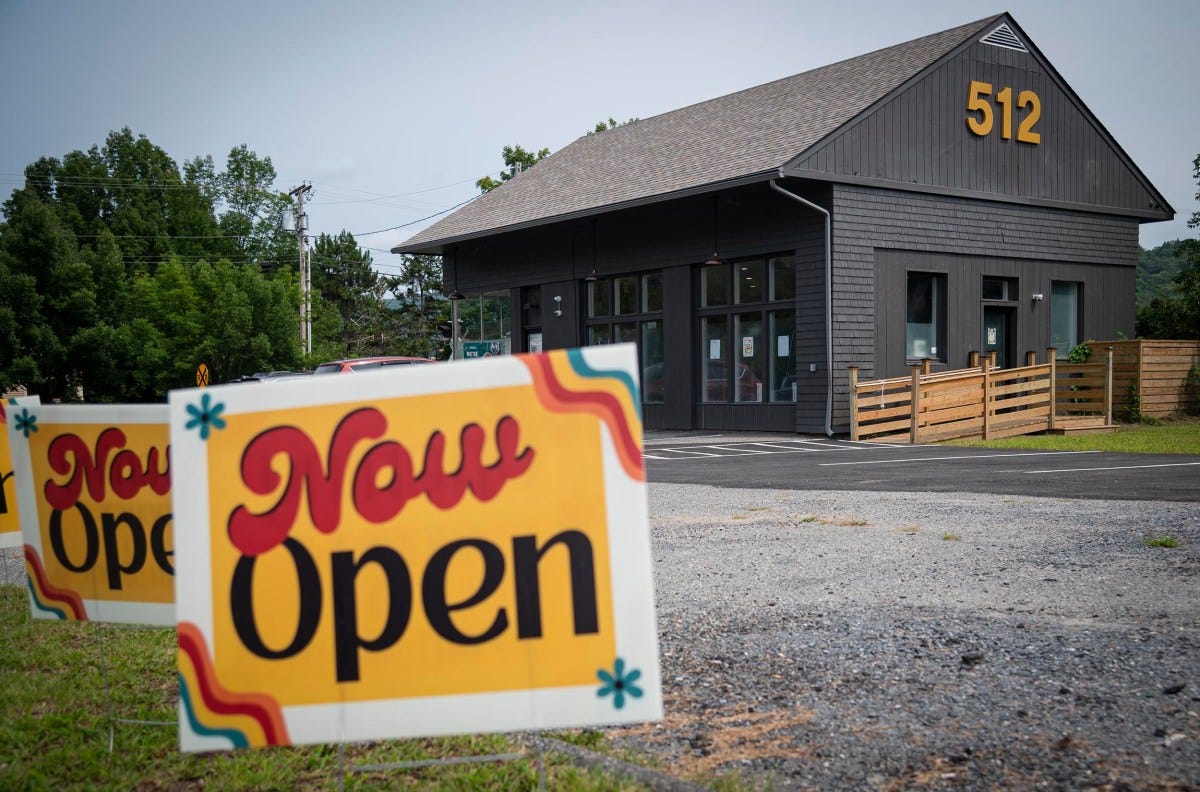 A grey building with the number 512 on it has colorful "Now Open" signs in the front yard. A grey building with the number 512 on it has colorful "Now Open" signs in the front yard.
