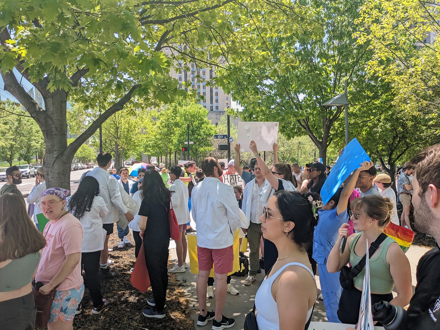 A large group of protestors, many wearing doctor's coats, congregated under a copse of trees.