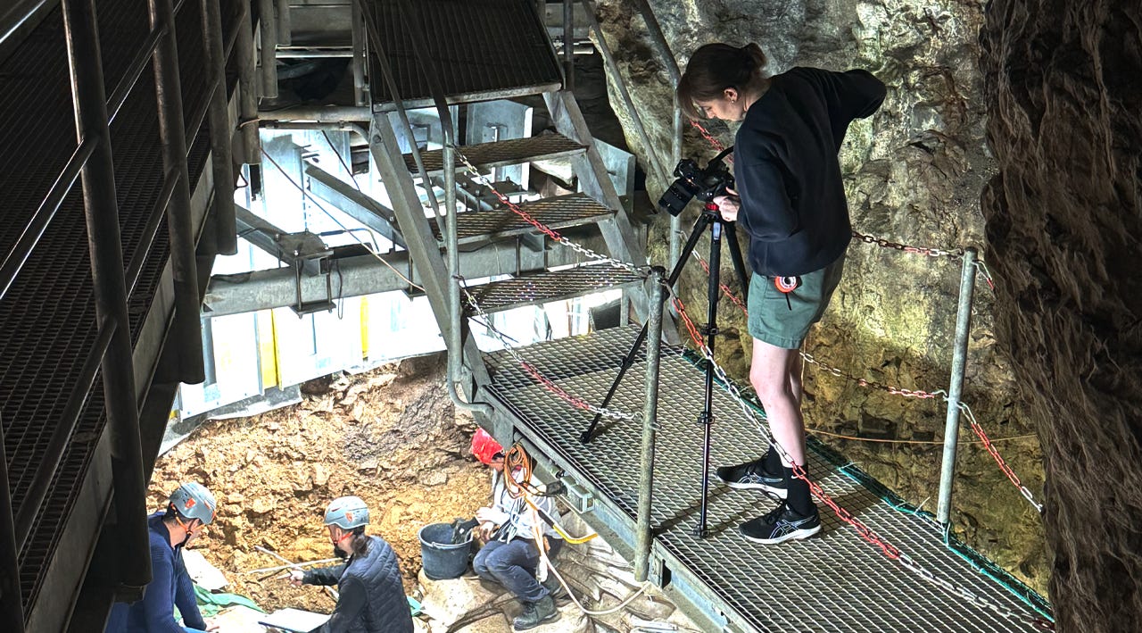 A woman in shorts stands on a walkway, filming archaeologists working on a dig site below her. A woman in shorts stands on a walkway, filming archaeologists working on a dig site below her.