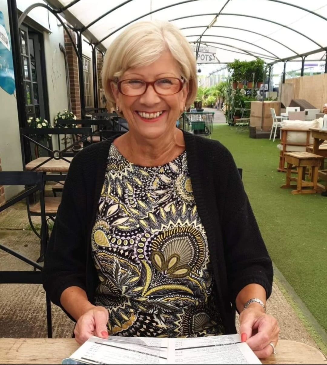 Picture of an older white lady with short blonde hair at glasses, sitting up a table and crying