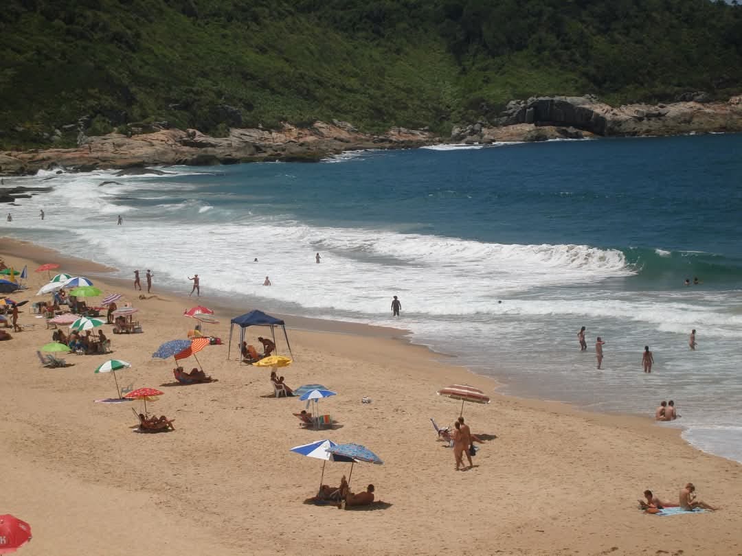 Wide view of Praia do Pinho showing a sandy beach bordered by green hills and rocky outcrops. People are scattered along the shore and in the water, some nude and others partially clothed, with umbrellas and small tents dotting the sand.