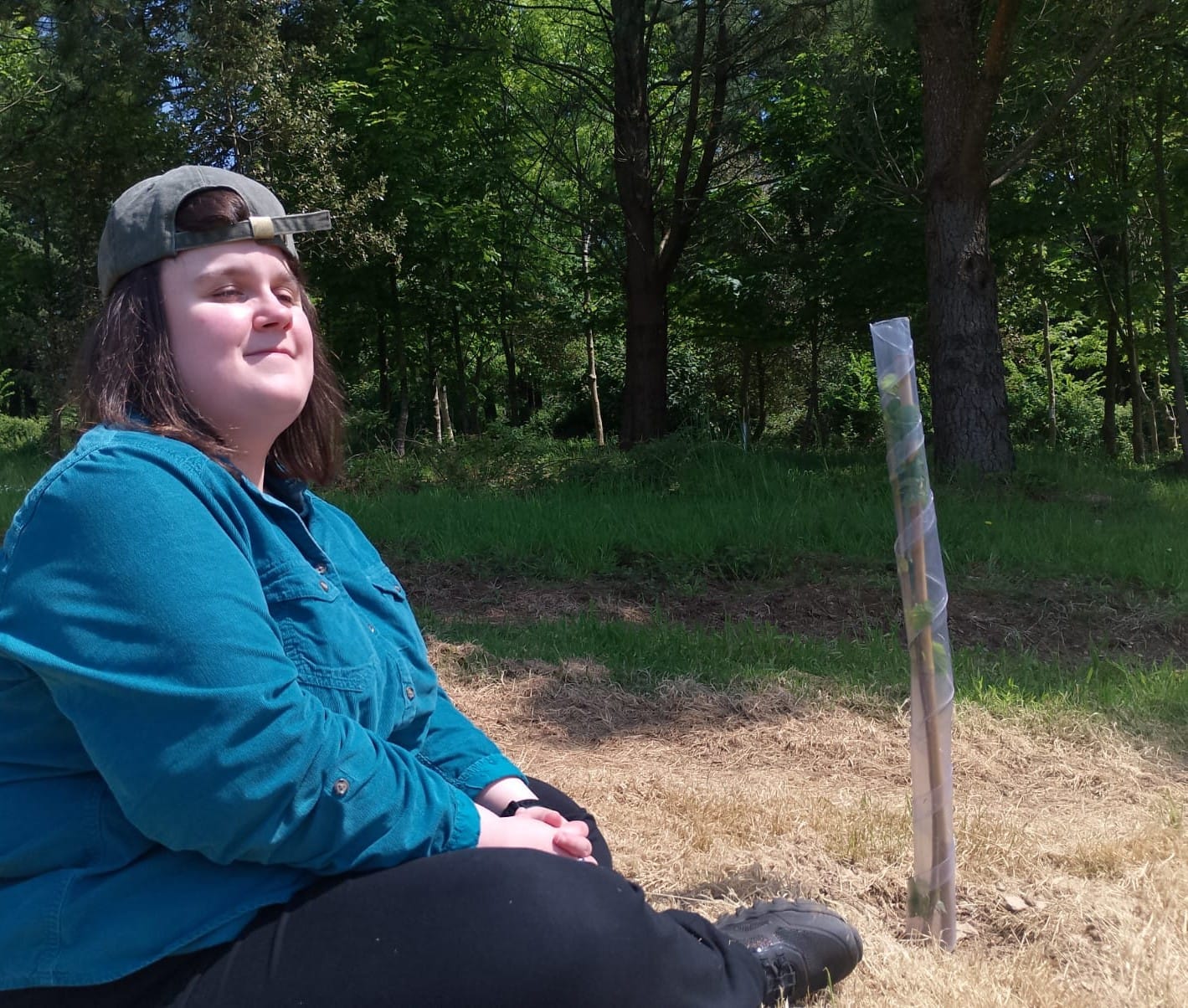 A young white woman wearing a green baseball cap and a teal shirt. She is sitting beside a sapling in a plastic guard.
