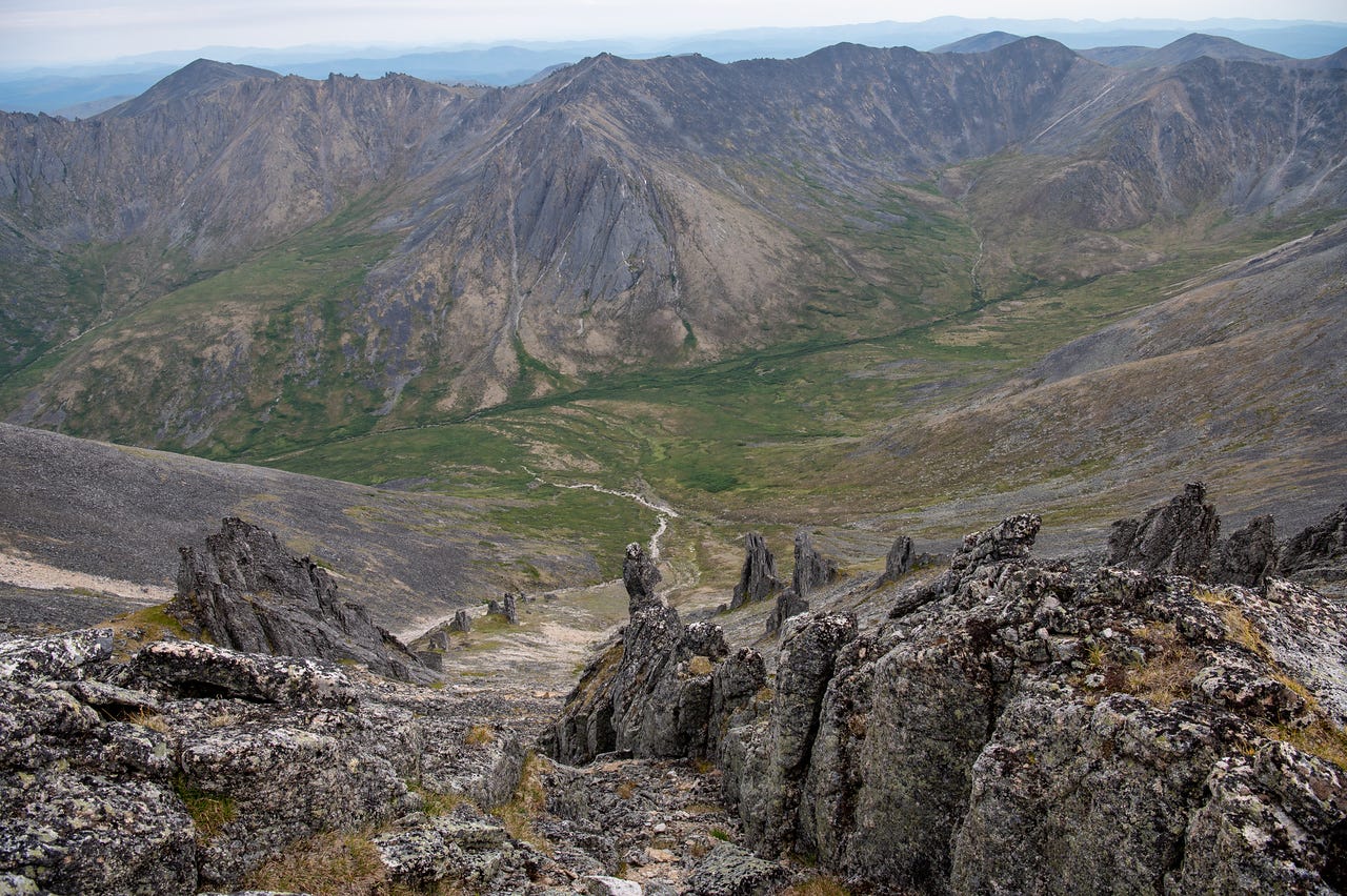A view down a long valley from among ragged granite tors in the foreground, the green valley floor and its thin stream winding between steep ridges toward far blue ranges under a pale sky.