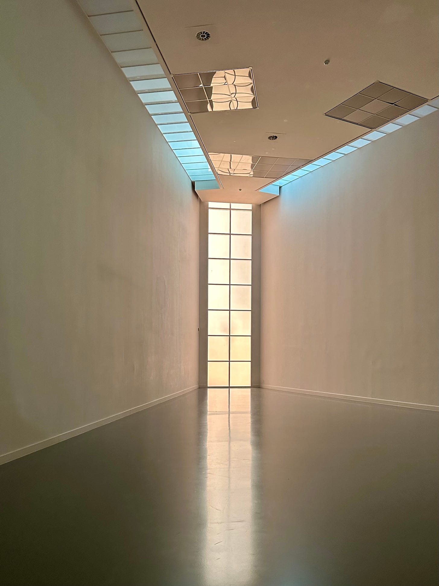 An empty gallery space. In this photo our gaze is directed toward a narrow vertical window stretching from floor to ceiling in this triangular space. There is a light-blue/pink haze shining through this room giving it an eery quality.