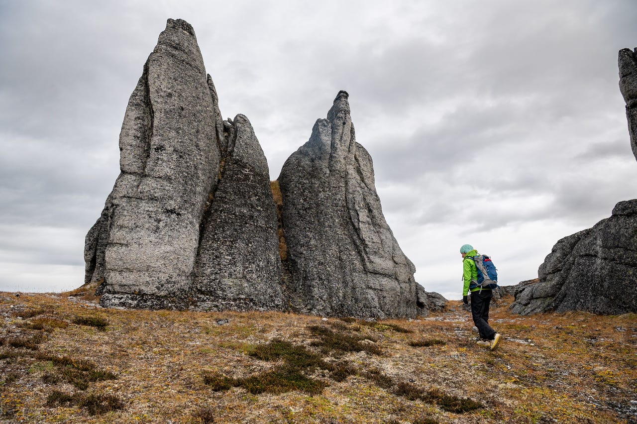 A hiker in a green jacket and blue hat walks toward massive granite tors known as the Windchimes at Mt. Prindle in the White Mountain National Recreation Area, Alaska. The tall gray rock formations rise vertically from the tundra, their weathered surfaces contrasting with the brown and green groundcover below. A cloudy gray sky looms overhead.