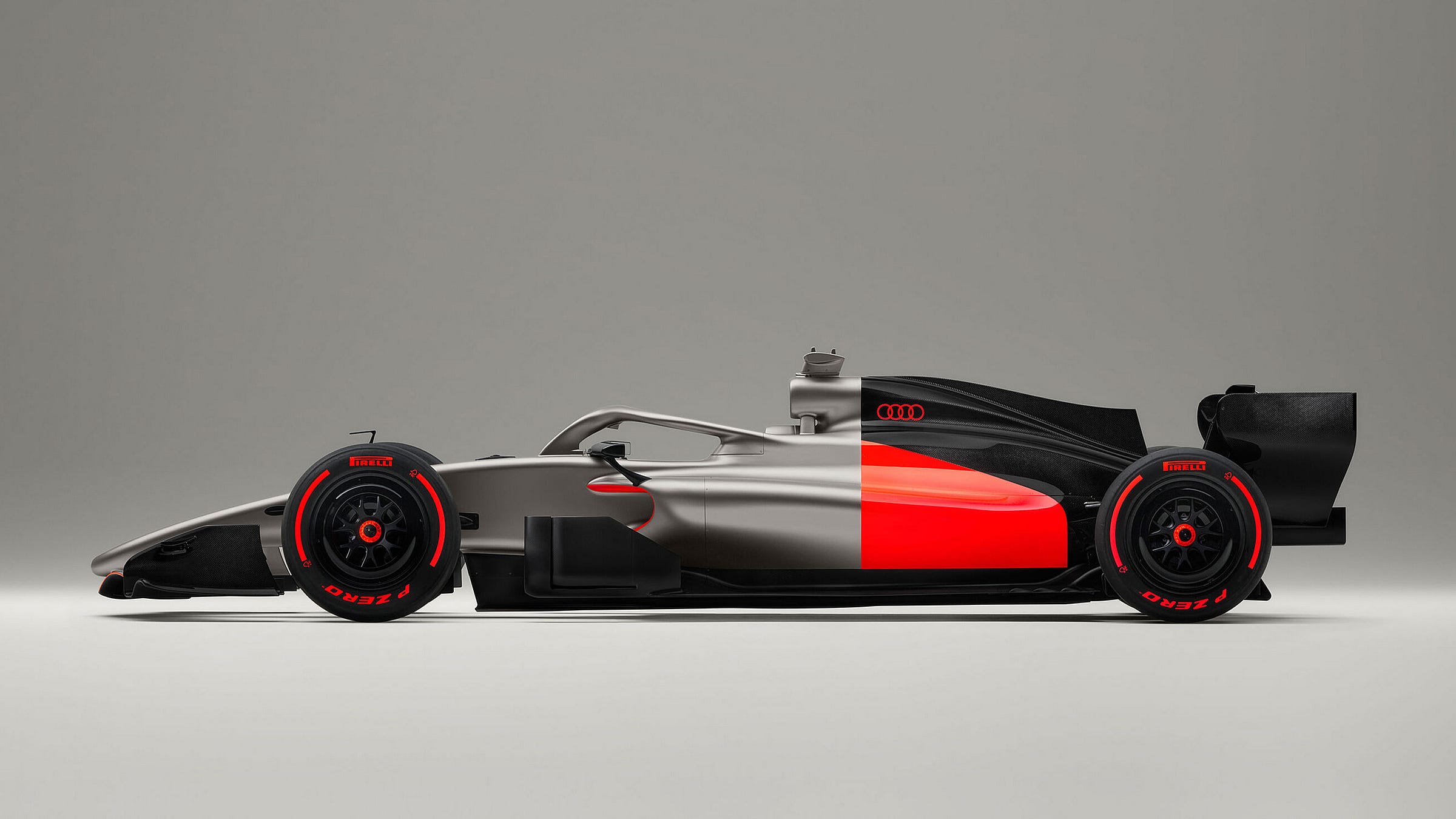 Side profile of the Audi R26 Formula One car against a light grey studio background. The livery combines matte silver on the nose and front section with a bold red sidepod panel and matte black engine cover and rear bodywork. The Audi four-ring logo is visible on the sidepod. Black multi-spoke wheels are fitted with Pirelli P Zero tyres with red lettering.