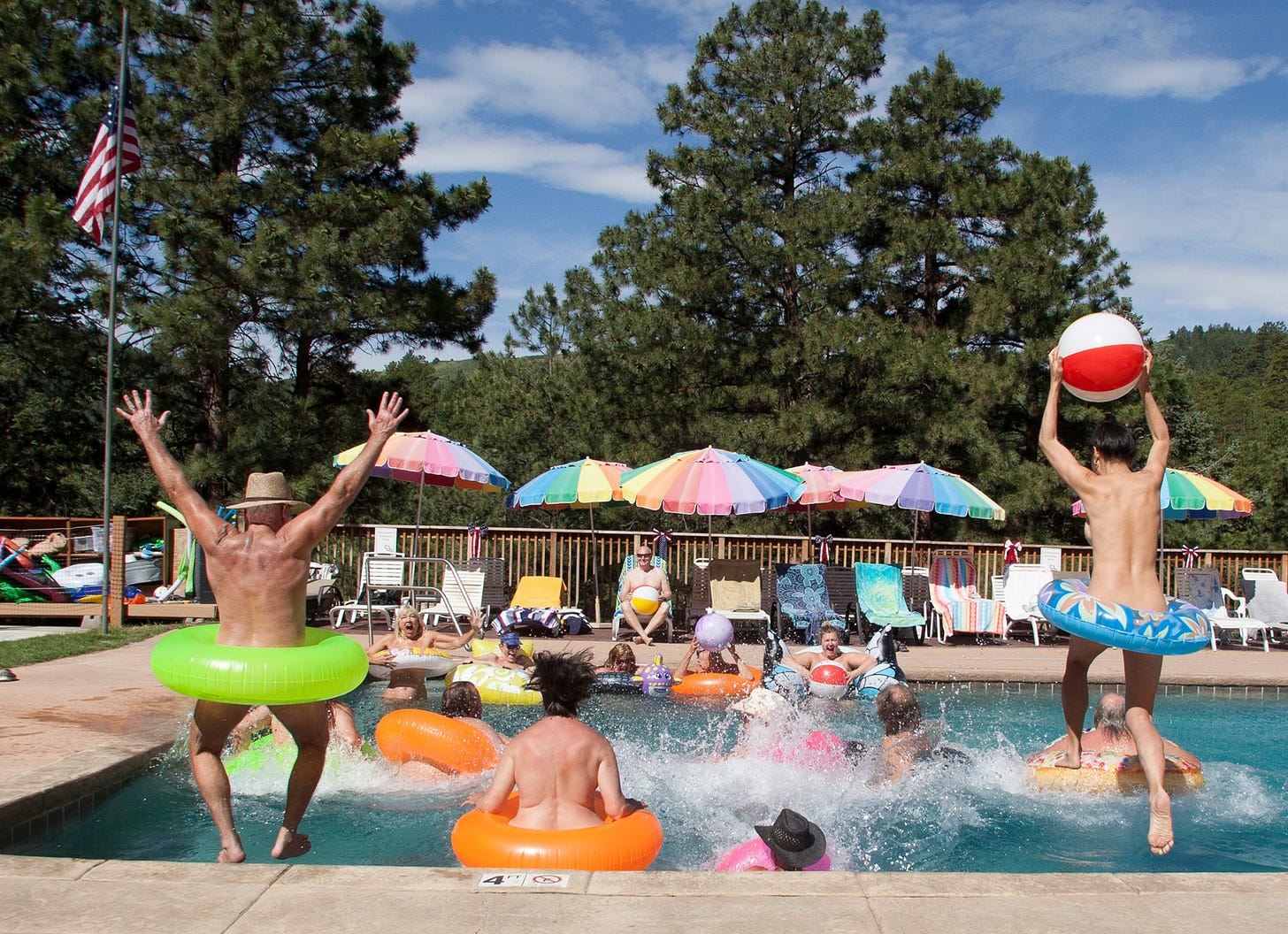 A lively pool scene at Mountain Air Ranch, showing a group of nude adults jumping and splashing into the water with inflatable tubes and beach balls. Two people leap midair from the pool’s edge—one with arms raised in a green float, the other holding a red-and-white beach ball. Rainbow-colored umbrellas and towels line the deck, with pine trees and blue sky in the background. © Marco Costick.