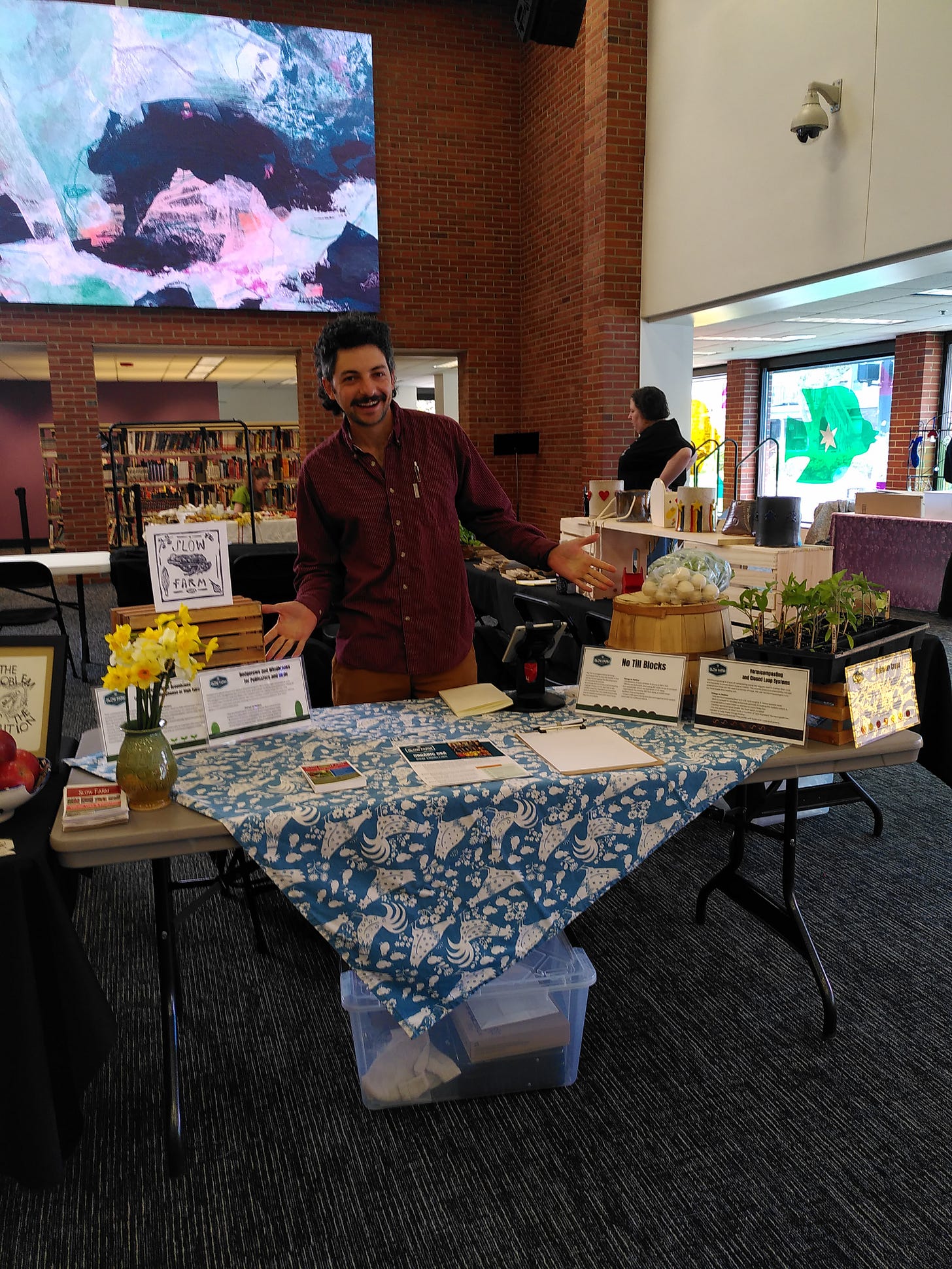 A mulleted man stands behind a table covered in signs plants and flowers.
