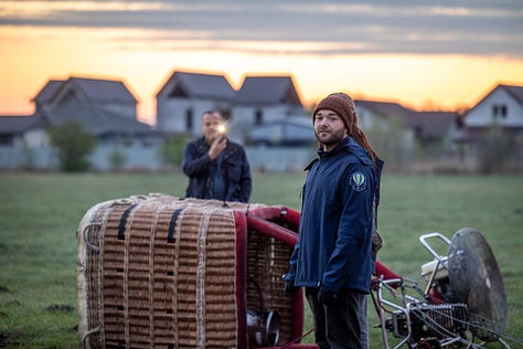 Înainte ca baloanele să urce, totul începe jos, pe iarbă, în frigul subțire al dimineții sau în lumina care abia se stinge peste câmp. Oamenii își ocupă locurile, verifică arzătoarele, întind pânza, ridică nacela, privesc cerul și, mai ales, ascultă vântul. În jur, mașinile stau cu farurile aprinse.