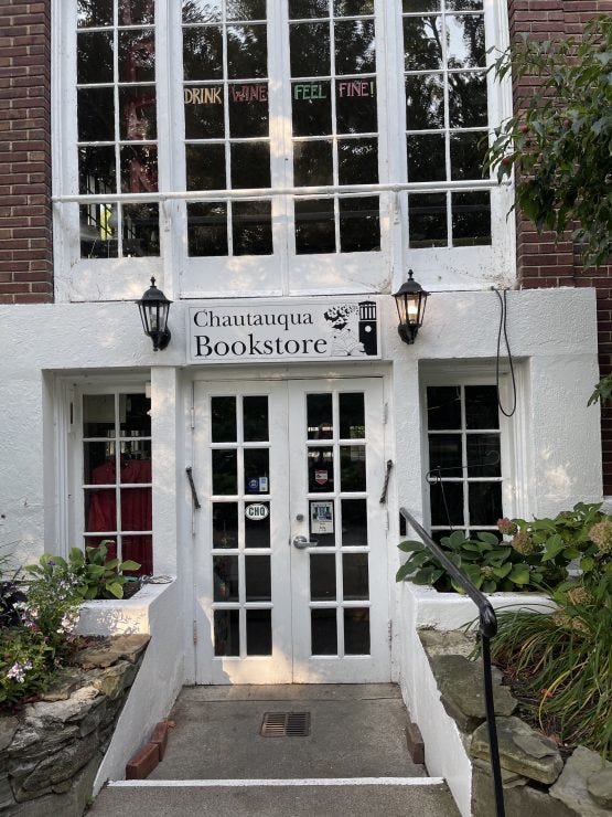 Storefront of the Chautauqua Bookstore featuring white French doors with multiple panes, flanked by black wall lanterns. A simple black and white sign reading 'Chautauqua Bookstore' sits above the entrance. Large windows above display a playful message 'DRINK WINE FEEL FINE!' The entrance is framed by brick walls, stone landscaping, and greenery