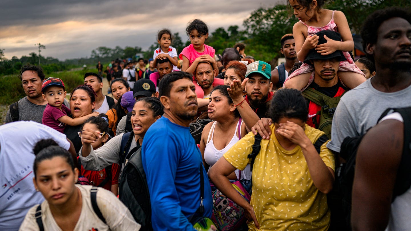 photo of crowded line of people wearing backpacks, several carrying a child on their shoulders, one woman with hand to forehead photo of crowded line of people wearing backpacks, several carrying a child on their shoulders, one woman with hand to forehead