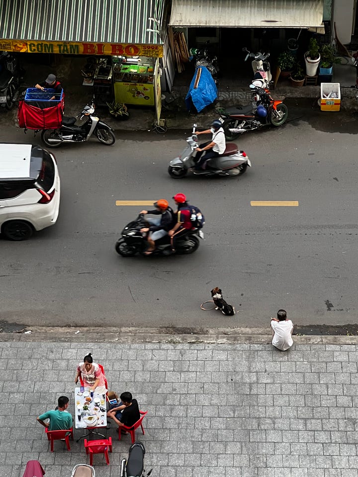 Daytime views of Ho Chi Minh City: a riverside skyline with high-rise buildings under dramatic clouds and construction along the riverbank, and a street scene with motorbikes passing a small “Com Tam Bi Bo” eatery while people dine on red plastic chairs and a dog sits near the curb.