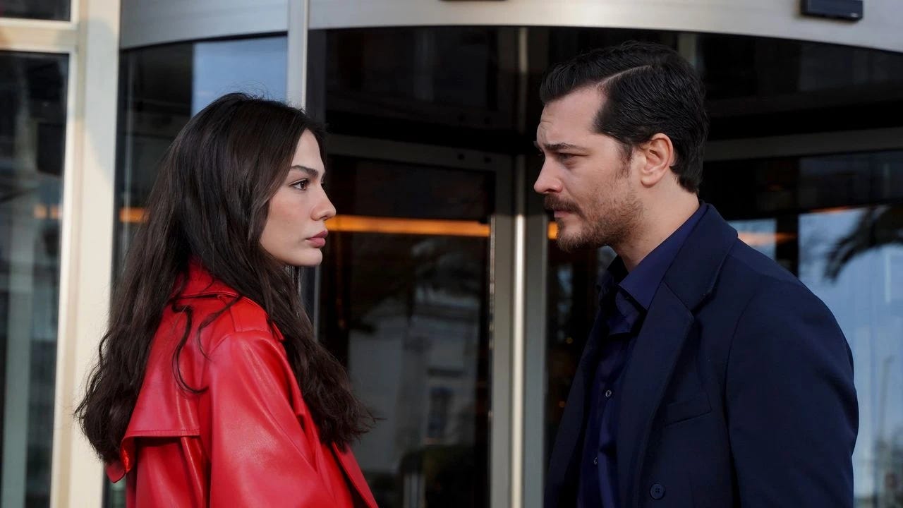 Bir diziden dramatik bir sahne: Parlak kırmızı deri ceket giyen bir kadın, cam kapıların önünde koyu mavi takım elbiseli bir erkekle yüz yüze duruyor. Bir diziden dramatik bir sahne: Parlak kırmızı deri ceket giyen bir kadın, cam kapıların önünde koyu mavi takım elbiseli bir erkekle yüz yüze duruyor.