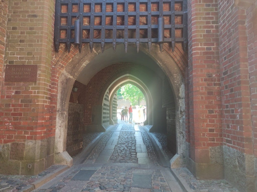 The walls are made of red brick. A high arch with raised portcullis leads into the courtyard, past two other gates. The ground is cobbled with large flat stones laid where cart wheels would roll