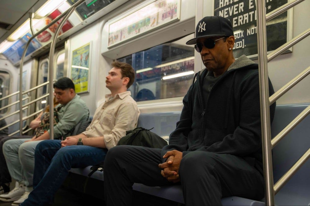 Denzel Wasington in dark cap and clothes sitting on the NYC subway in Highest 2 Lowest