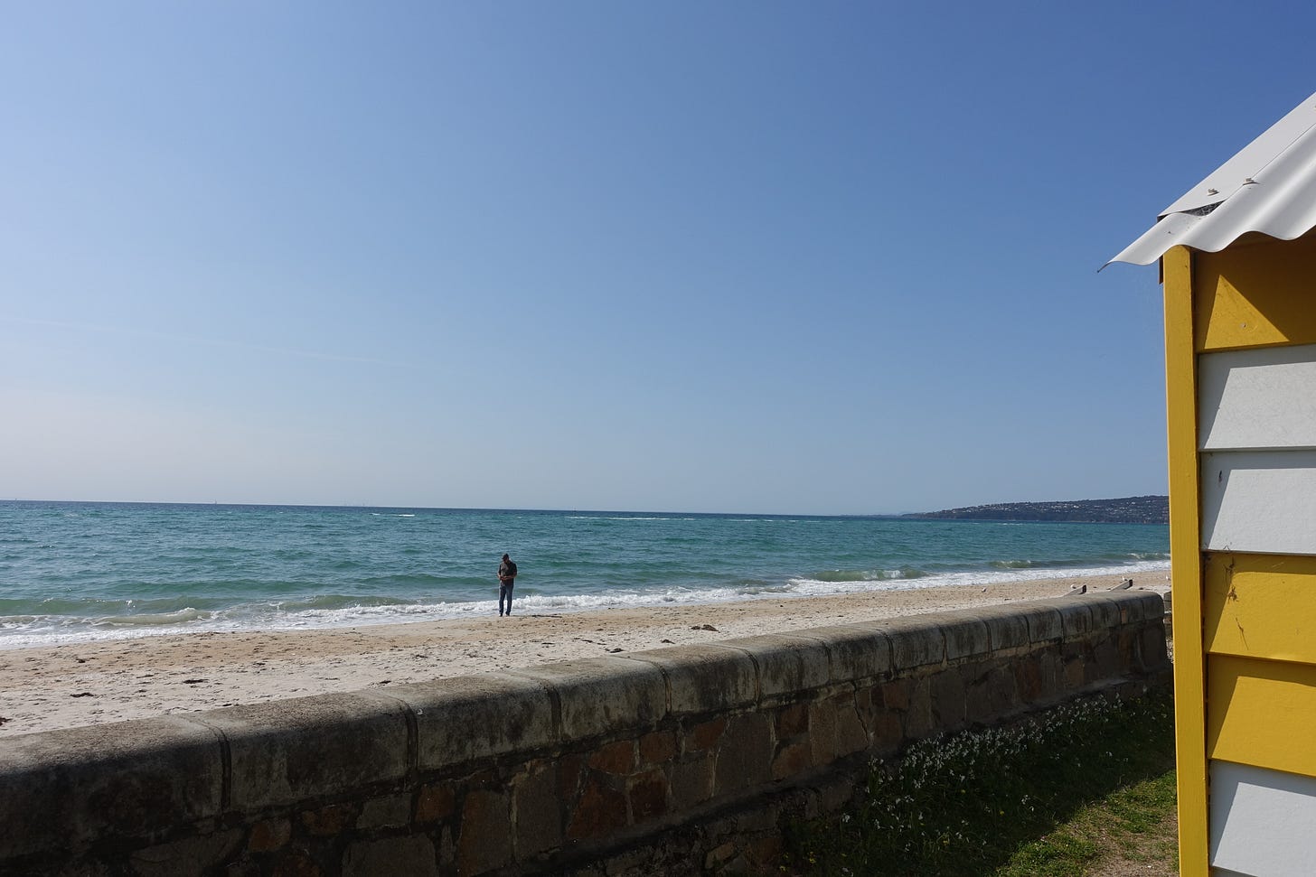 Beach scene with stone seawall, yellow and white beach box visible on right, and solitary figure standing at water's edge against blue ocean horizon
