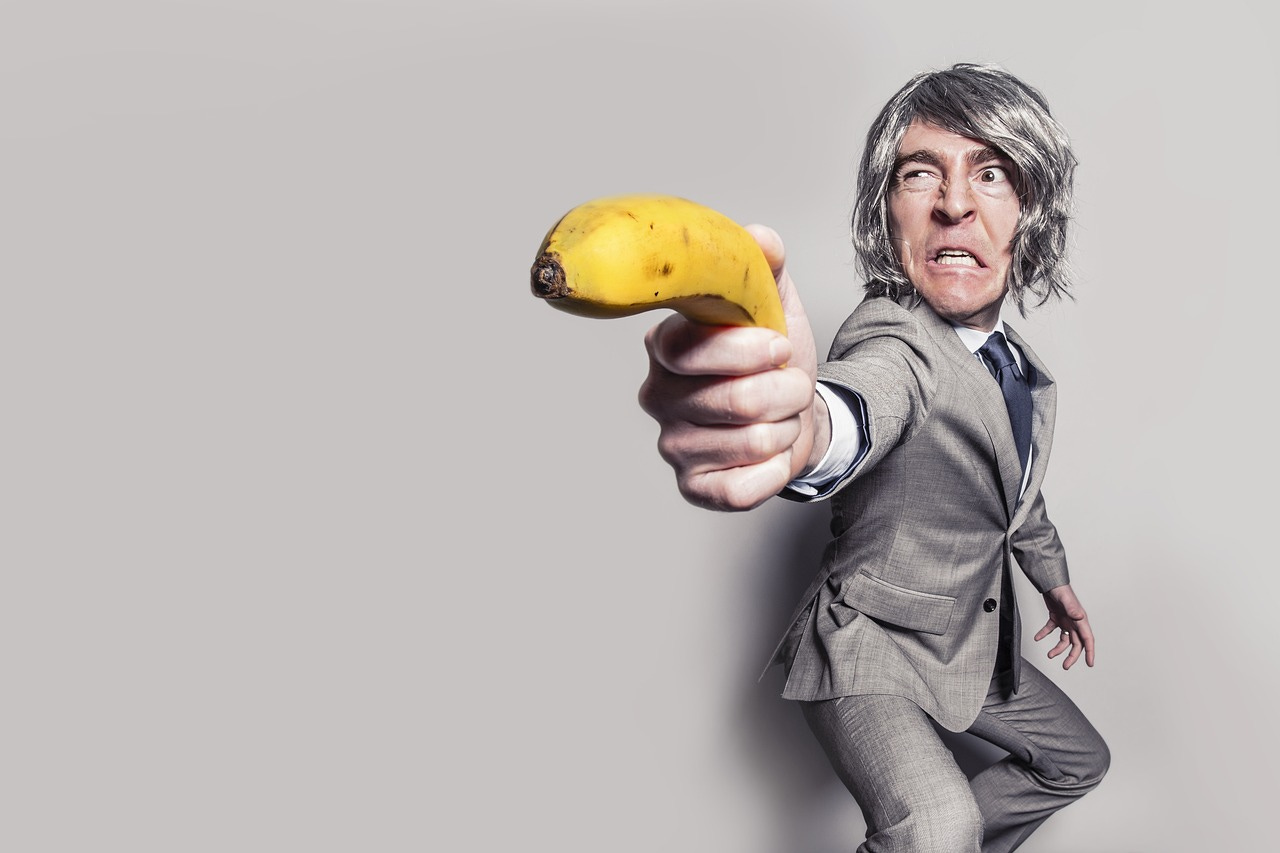 Photo of older man in heather grey suit and dark grey tie squatting in a defensive martial arts posture, long dark grey hair to his chin. He has a angry look in his face and he holds a yellow banana like it is a gun 