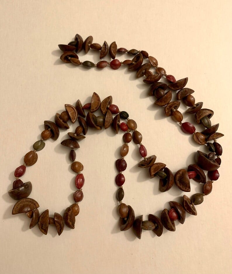 May include: A handcrafted necklace featuring a unique design with brown, red, and green seed beads. The necklace is composed of various shapes and sizes of natural seeds, creating a rustic and earthy aesthetic. The beads are strung together, forming a long, elegant piece of jewelry.