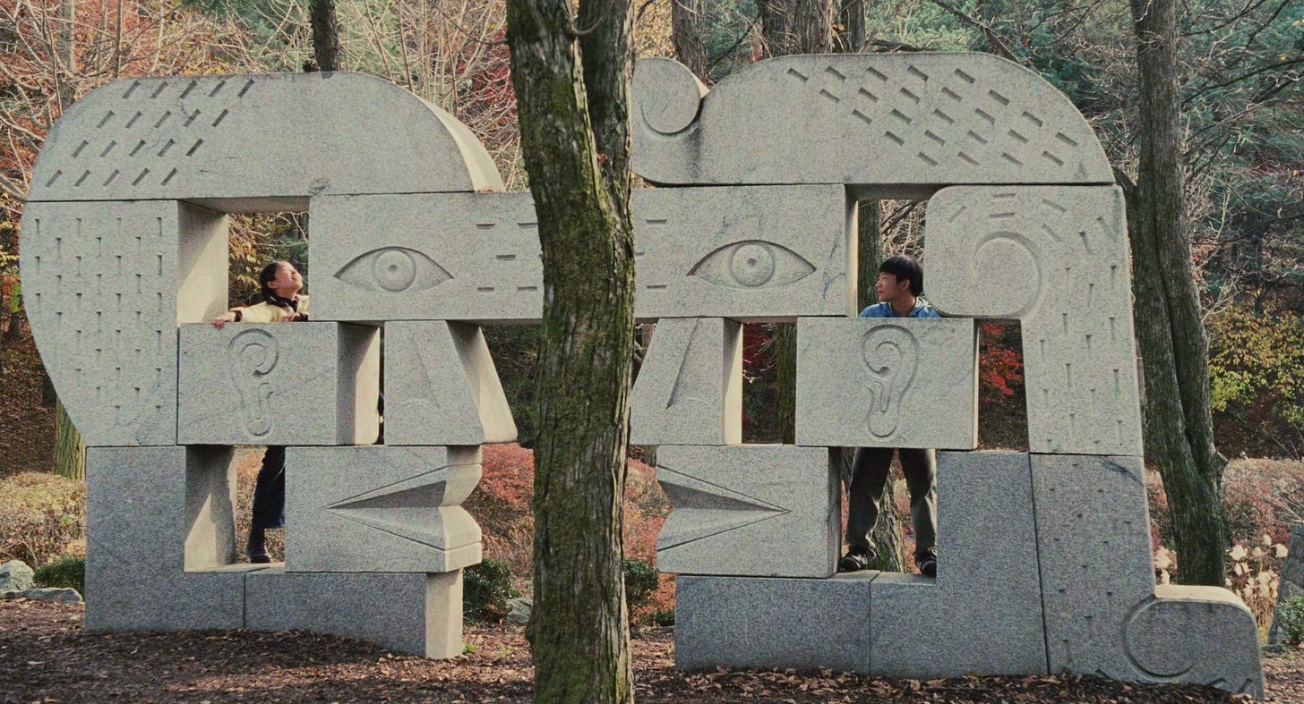 Dois jovens olhando um para o outro através de uma escultura de pedra em formato de faces estilizadas em um parque, representando a conexão e a distância entre Hae Sung e Na Young/Nora.