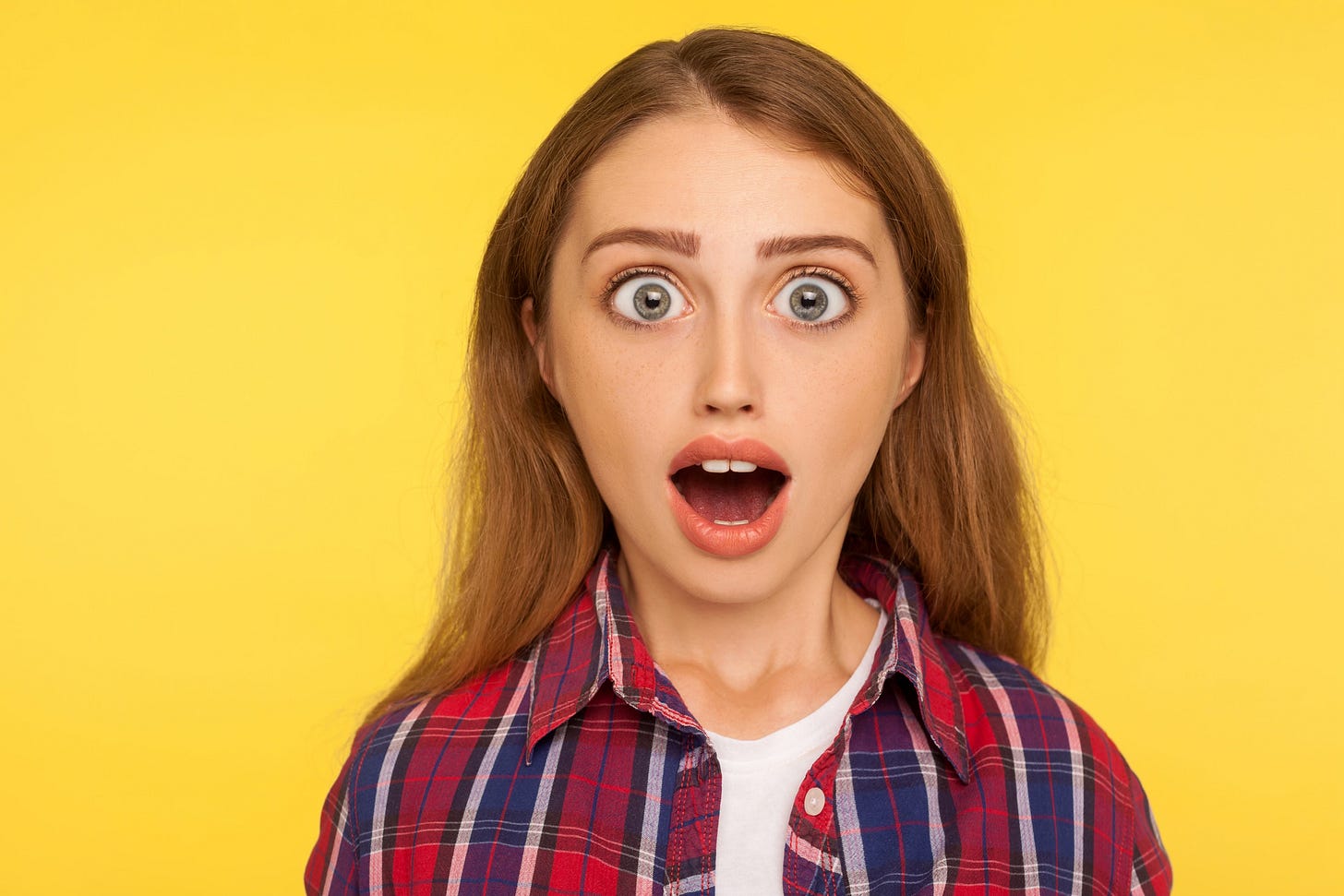 The image shows a young woman with long brown hair wearing a red, blue, and white plaid shirt over a white t-shirt. She has an expression of surprise or shock on her face, with her eyes wide open and mouth agape. The background is solid yellow, emphasizing her expression. The image shows a young woman with long brown hair wearing a red, blue, and white plaid shirt over a white t-shirt. She has an expression of surprise or shock on her face, with her eyes wide open and mouth agape. The background is solid yellow, emphasizing her expression.