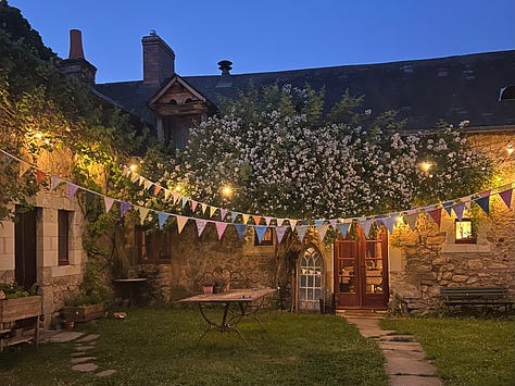 A collage of the grounds around the rented farmhouse, including an iron gate set into an ivy-covered stone wall, a garden path, a courtyard strung with bunting at dusk, two small children watching a rainbow from a doorway, pink cyclamen carpeting the ground beneath a large tree, and children walking down tree-lined country paths.