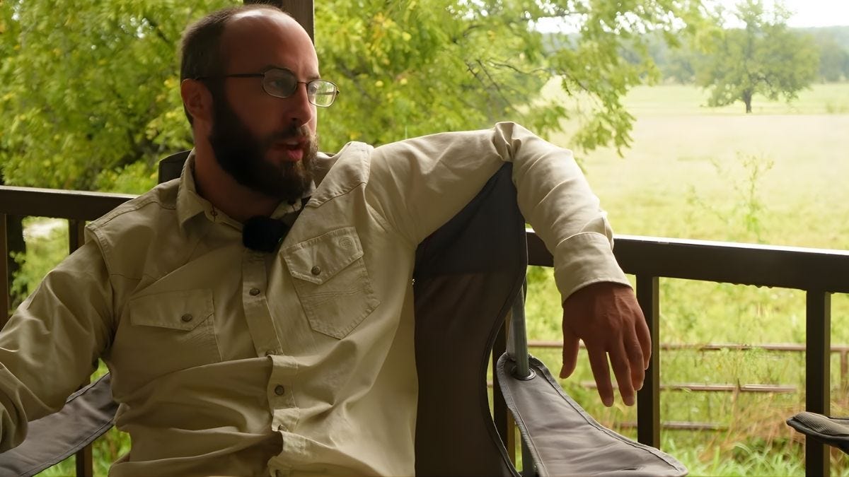 A rancher in a khaki work shirt sits on a porch overlooking green pastures, speaking during an interview about regenerative ranching and rural renewal.