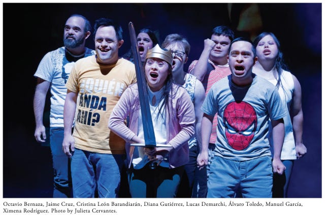 An ensemble of actors with Down syndrome stands together, their mouths open and fists clenched. A woman at the front of the group, wearing a crown, holds a sword. An ensemble of actors with Down syndrome stands together, their mouths open and fists clenched. A woman at the front of the group, wearing a crown, holds a sword.