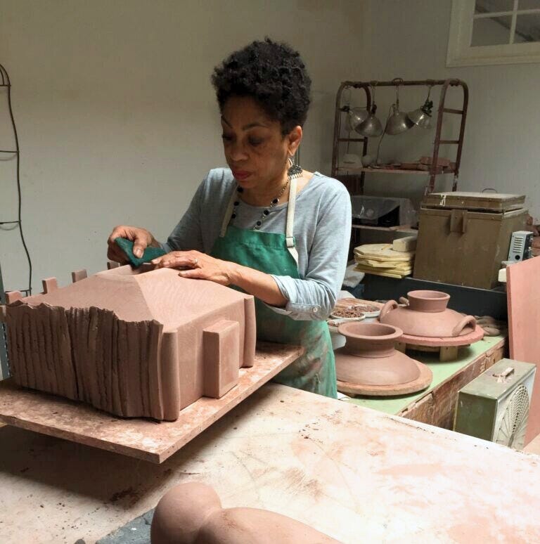A Black woman wearing a clay-smudged apron shapes a large piece of clay in her pottery studio, surrounded by ceramic tools and various clayworks in progress. 