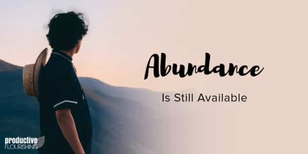 A man stands facing a valley with his back to the camera, a straw hat hangs to the back from his neck. Text Overlay: Abundance is Still Available. A man stands facing a valley with his back to the camera, a straw hat hangs to the back from his neck. Text Overlay: Abundance is Still Available.