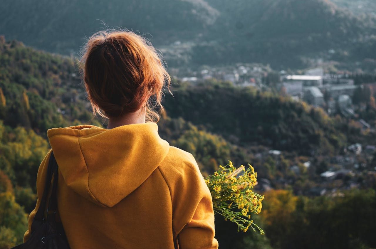 Back view of a woman in a mustard colored hoodie with red hair holding yellow flowers and looking out at the surrounding hills.