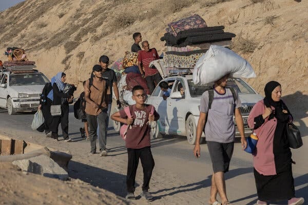 Palestinians of varying ages are carrying their belongings as they walk on foot. Behind them is a car piled high with mattresses. 