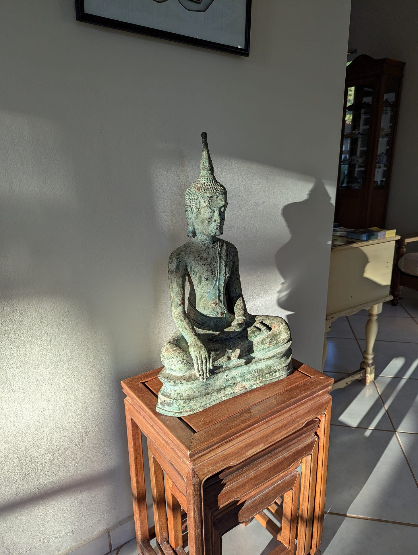 Photo of a Buddhist statue in a guesthouse reception area, with light and shadow cast on it. The statue has one hand pointing down to the ground and sits on a small wood table.