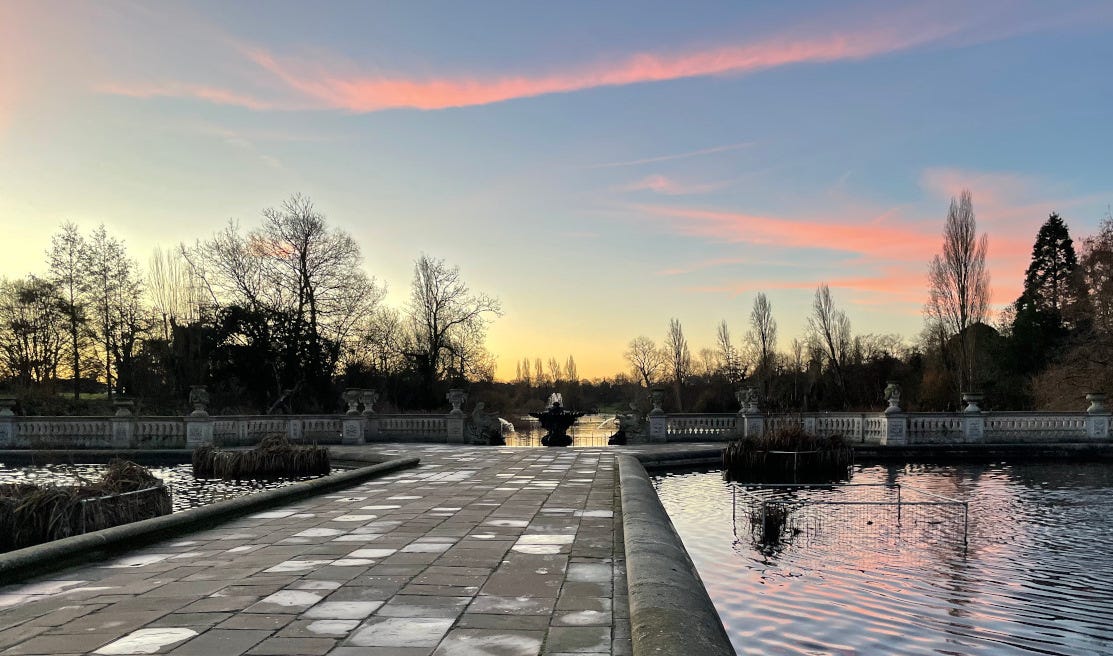 view of London's Italian Gardens at dawn, with sky fading from yellow to blue with pink streaks, trees in silhouette, water on either side of paved walkway and Serpentine in background