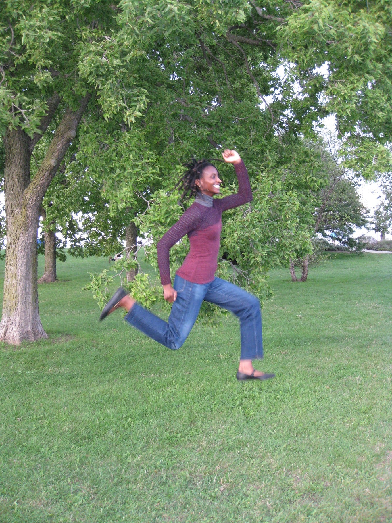 An image of myself in the same outfit as before (a stripped top with mesh sleeve and a grey turtleneck underneath, dark denim, and black ballet flats.) I am jumping, recreating the Super Mario pose with one arm up and the other down, my legs in a mid-air march.