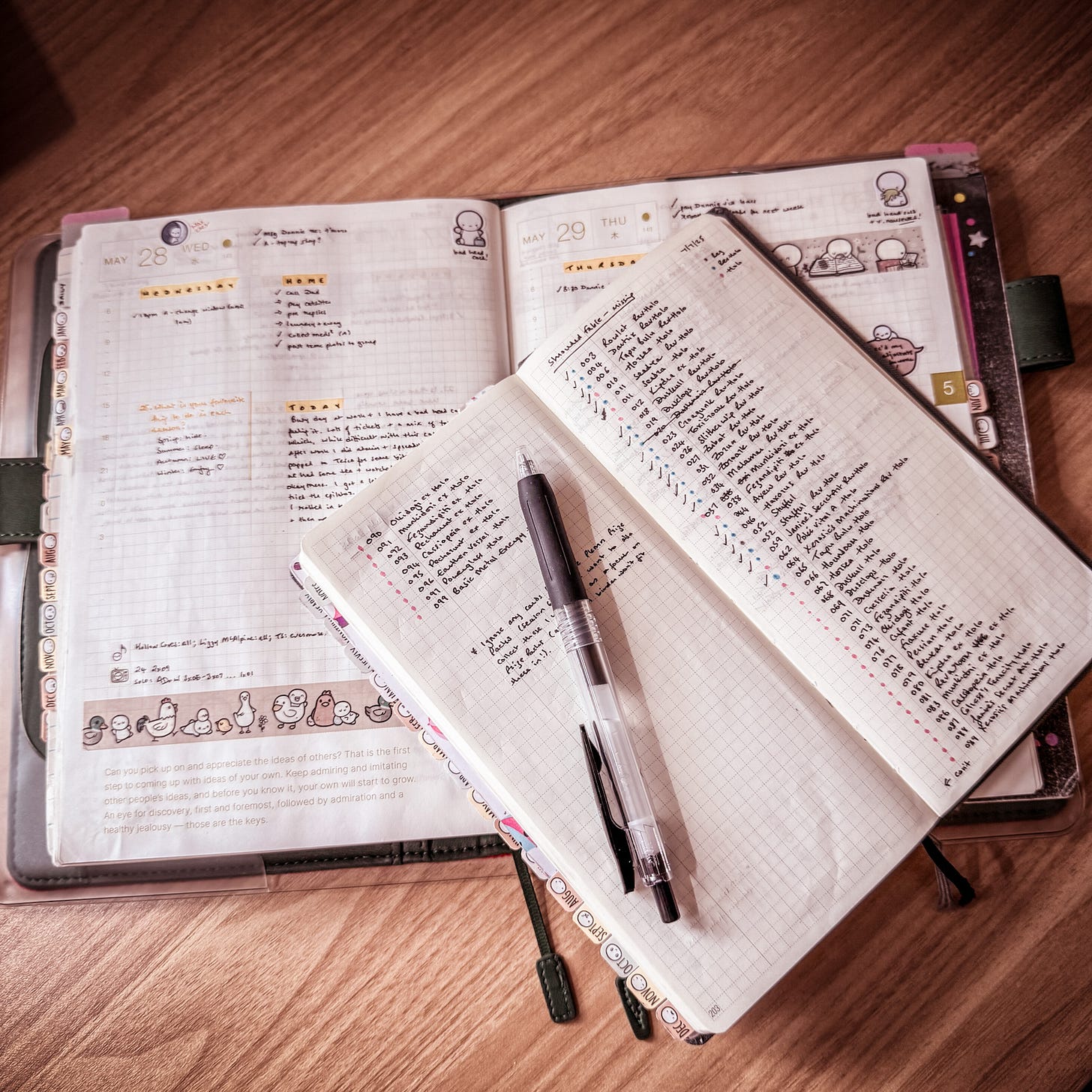 An open Hobonichi A5 Cousin planner is shown on a wooden desk, its pages lying flat and showing some planner entries. An open Hobonichi Weeks lies slightly on top of it, showing a two-page spread of notes in black ink. A black gel pen lies on top of one of its pages.
