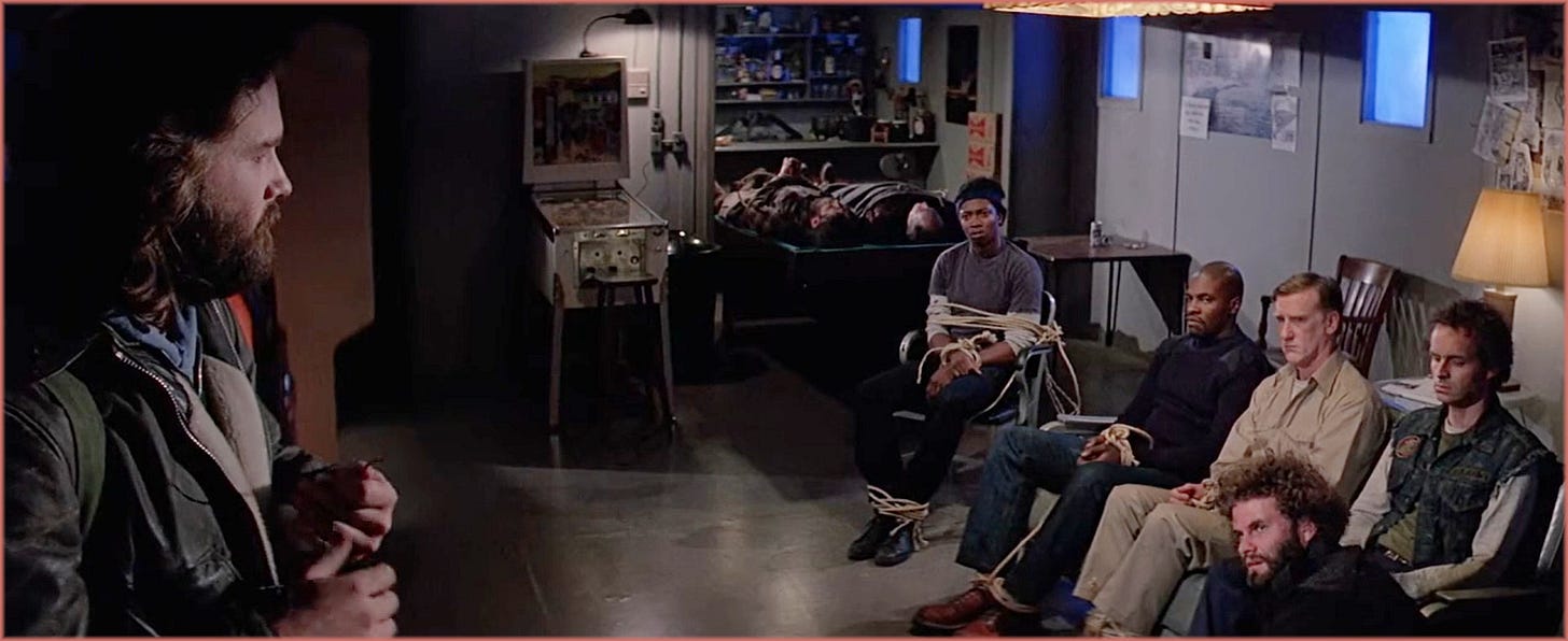 Wide shot from The Thing (1982) blood test sequence: MacReady in profile on the left, five men seated and tied to chairs on the right, two bodies visible on tables in the background. Carpenter's anamorphic frame keeps every living suspect and every dead one simultaneously in view.
