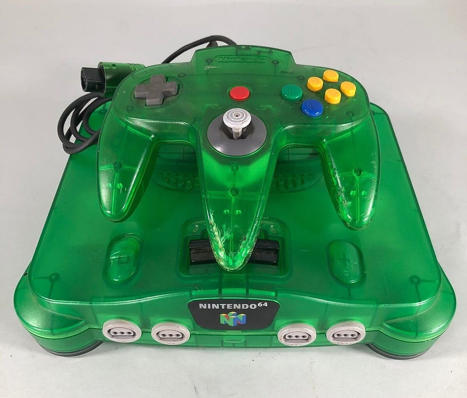 Green Nintendo 64 console and controller set on a light surface. Transparent casing with colorful buttons, brand logo visible.