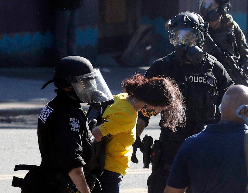 caption: Police arrest a person wearing a yellow shirt an armband that reads "Mom," Saturday, July 25, 2020, during a Black Lives Matter protest near Seattle Central Community College in Seattle. A large group of protesters were marching Saturday in Seattle in support of Black Lives Matter and against police brutality and racial injustice.