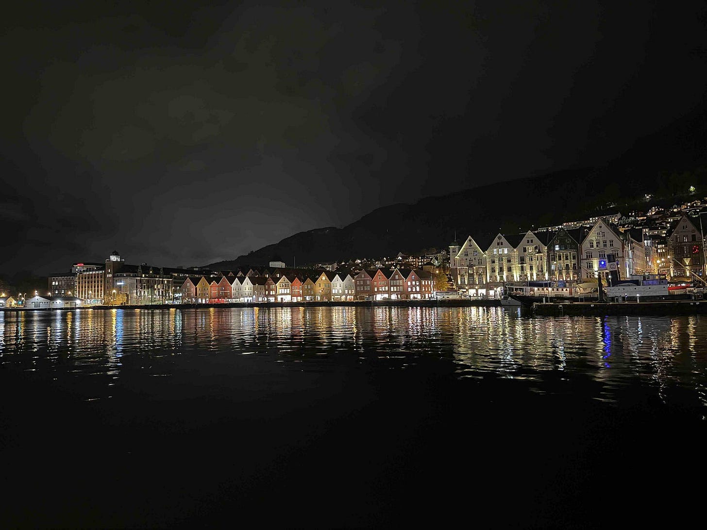 A nighttime waterfront view of Bryggen in Bergen, Norway, featuring a row of brightly lit, colorful wooden buildings with steep gabled roofs. Their reflections shimmer on the calm water in front. Behind the historic harbor district, a hillside dotted with lights from homes rises into the dark sky, adding depth to the scenic and culturally rich landscape.